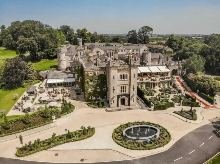 An aerial perspective of Cabra Castle Hotel showing the expansive Neo-Gothic stone architecture, formal gardens with a circular fountain, and the grand red-carpeted entrance staircase.