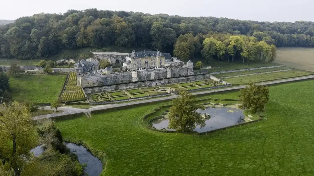 An elevated aerial view of Château Neercanne in Maastricht, showcasing its tiered yellow marlstone architecture, symmetrical Baroque gardens, and territorial position overlooking the Jeker Valley.