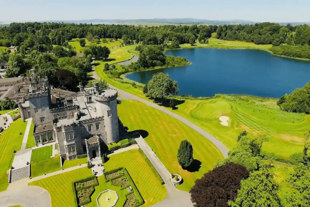 The 19th-century Neo-Gothic limestone facade of Dromoland Castle featuring its dramatic crenelated battlements and original baronial stonework mirrored in the 24-acre Lough Dromoland lake view.