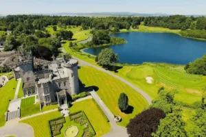 The 19th-century Neo-Gothic limestone facade of Dromoland Castle featuring its dramatic crenelated battlements and original baronial stonework mirrored in the 24-acre Lough Dromoland lake view.