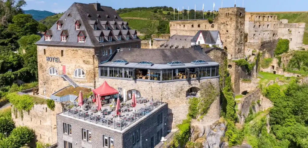 An aerial view of Hotel Schloss Rheinfels integrated into the historic stone ruins of Rheinfels Castle, featuring a large outdoor terrace and a modern glass-enclosed restaurant overlooking the Rhine Valley.