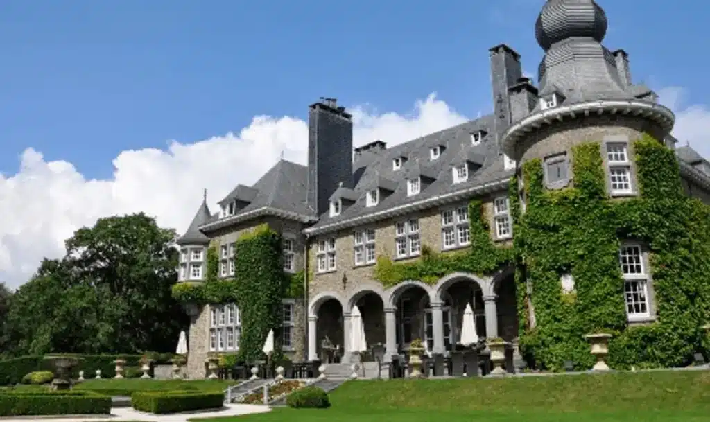 The stone facade of Manoir de Lébioles in the Belgian Ardennes, featuring ivy-covered turrets, a slate roof with dormer windows, and a formal stone terrace overlooking manicured gardens.