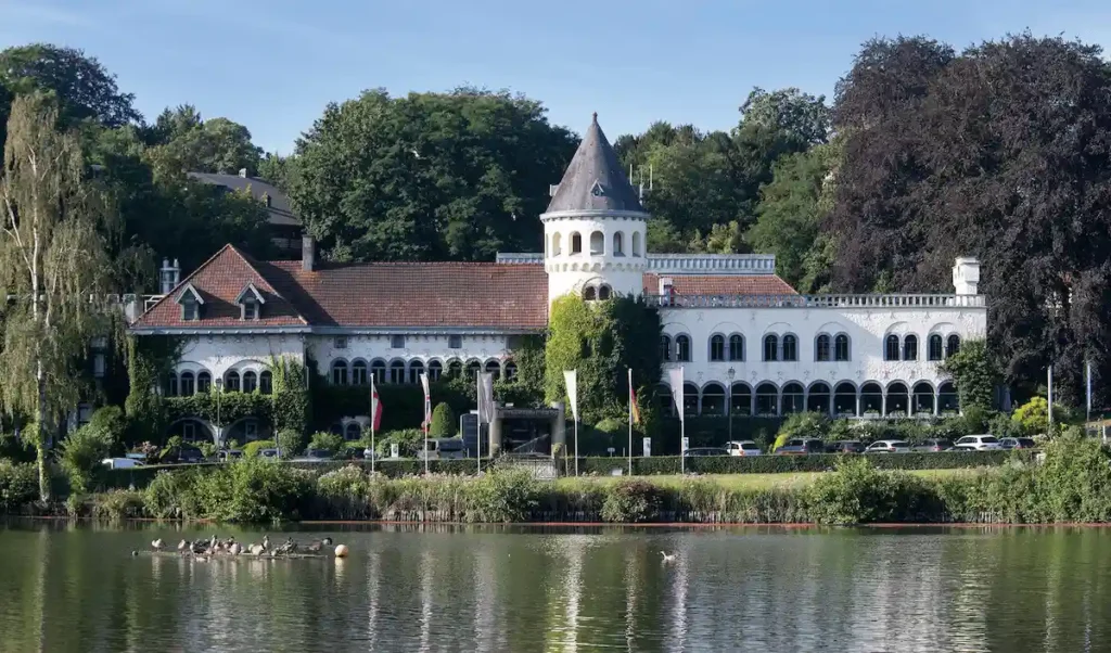 The white-walled facade of Martin's Château du Lac in Genval, featuring a central ivy-clad turret, red-tiled roofing, and arched windows overlooking the tranquil lake and surrounding greenery.