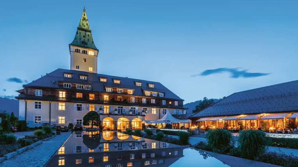 The illuminated facade of Schloss Elmau at dusk, featuring its iconic green-roofed clock tower and modern reflective pool set against the silhouette of the Bavarian Alps.