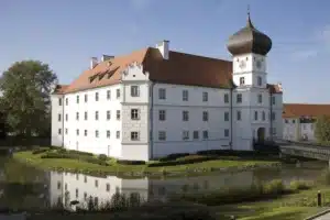 The pristine white facade of Schloss Hohenkammer in Bavaria, featuring a distinctive onion-domed clock tower and red-tiled roof, reflected in the calm waters of its surrounding moat.