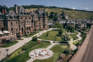 An elevated view of the grand Neo-Renaissance facade of Schloss Lieser along the Mosel River, featuring ornate stone carvings, slate-roofed spires, and a circular courtyard set against terraced vineyards.
