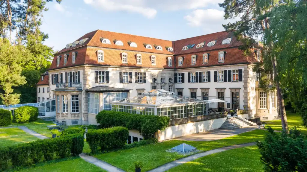 The garden-side view of Schlosshotel Berlin, showcasing the Neo-Renaissance architecture, a large glass-enclosed conservatory, and manicured lawns surrounded by the private greenery of the Grunewald forest.