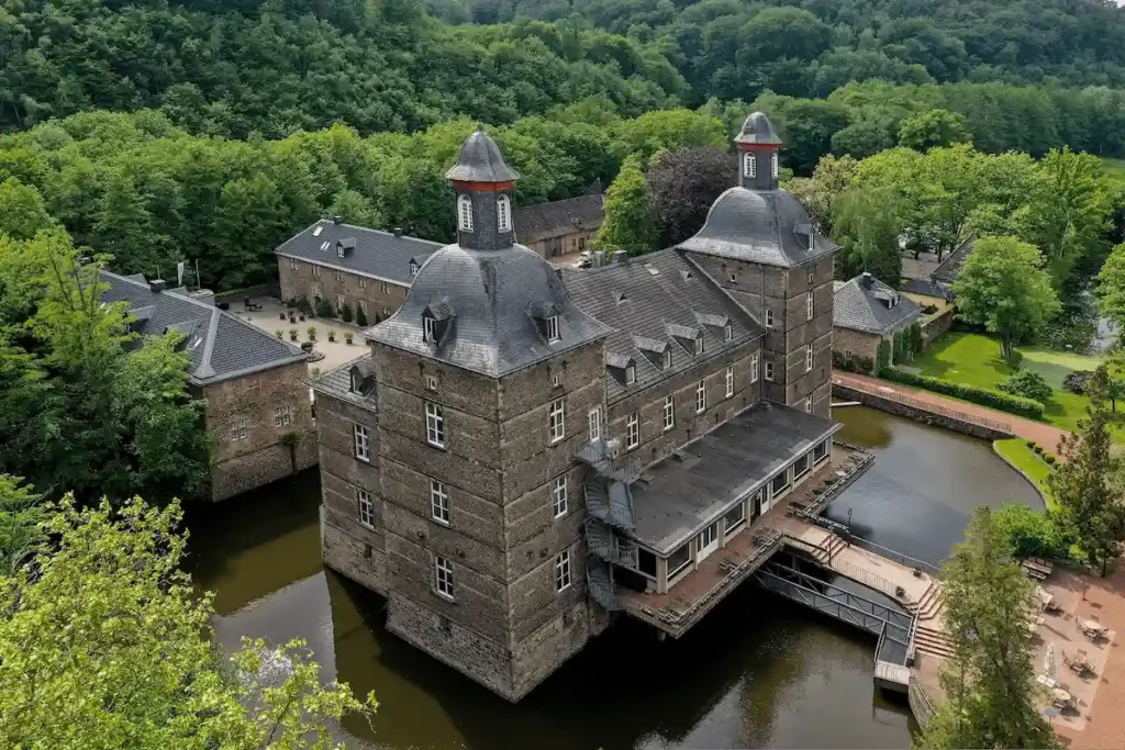 An aerial perspective of the dark stone Schlosshotel Hugenpoet in Essen, a 17th-century moated castle featuring prominent corner turrets with slate domes, set within a lush forest and reflected in its surrounding defensive waters.