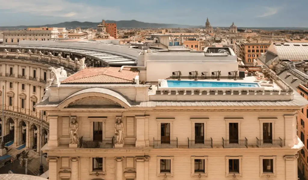 An aerial view of the Anantara Palazzo Naiadi Rome Hotel, showing the rooftop terrace and infinity pool overlooking the neoclassical architecture of Piazza della Repubblica and the Roman skyline.