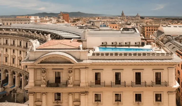 An aerial view of the Anantara Palazzo Naiadi Rome Hotel, showing the rooftop terrace and infinity pool overlooking the neoclassical architecture of Piazza della Repubblica and the Roman skyline.