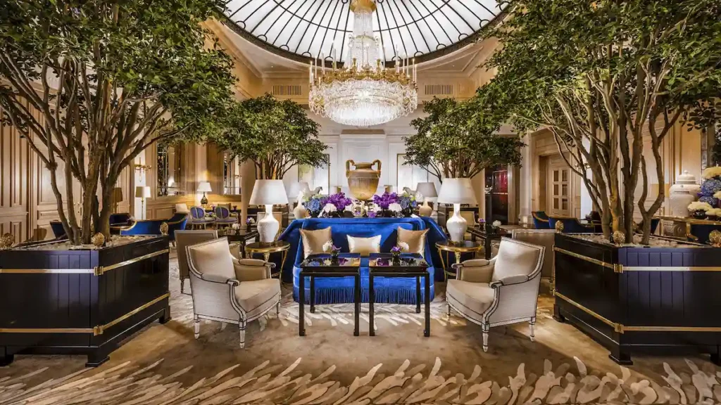 The iconic "Le Hall" lobby at Baur au Lac, featuring indoor trees, plush blue velvet seating, and a grand crystal chandelier beneath a glass dome.