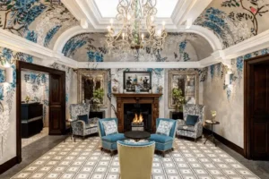 The light-filled reception lounge of Brown's Hotel London featuring vaulted white ceilings adorned with hand-painted blue wisteria murals, a grand crystal chandelier, and a traditional wood-burning fireplace surrounded by teal velvet wingback chairs.