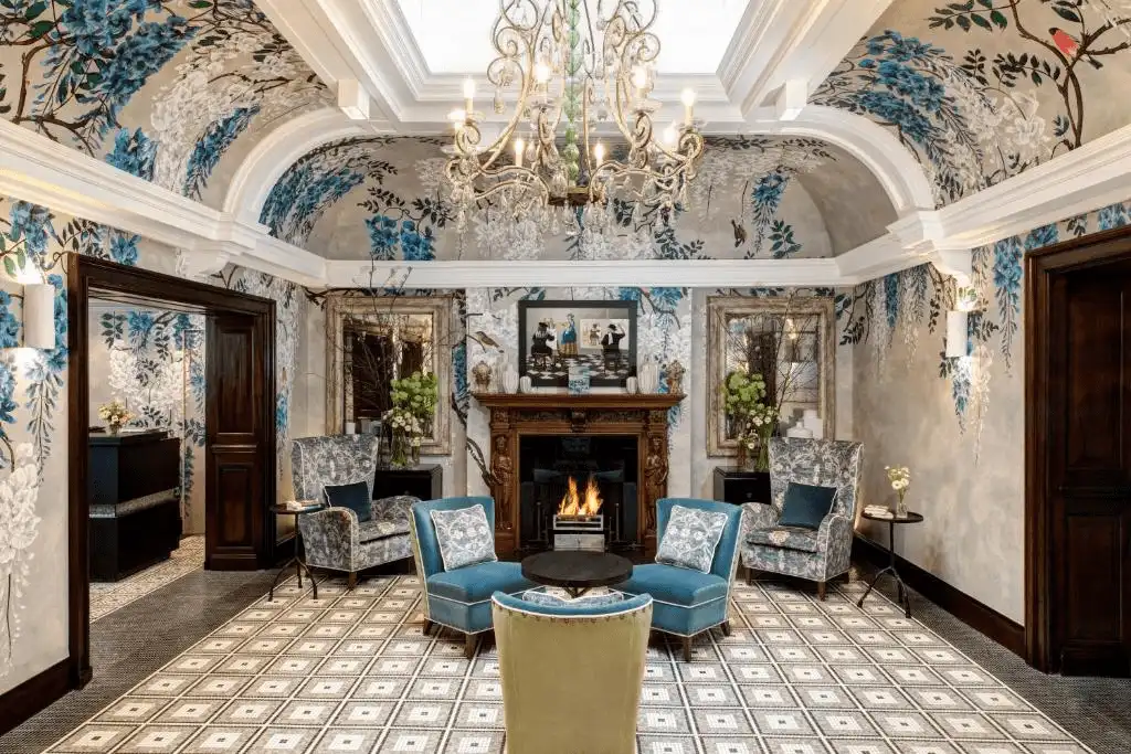 The light-filled reception lounge of Brown's Hotel London featuring vaulted white ceilings adorned with hand-painted blue wisteria murals, a grand crystal chandelier, and a traditional wood-burning fireplace surrounded by teal velvet wingback chairs.