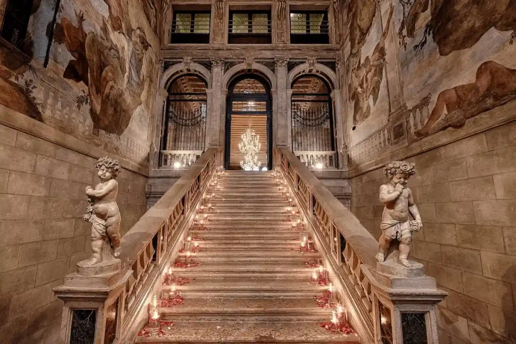 The 1732 "Scalone dei Giganti" (Staircase of the Giants) by Andrea Tirali at Ca' Sagredo Hotel Venice, featuring marble cherub sculptures and expansive 18th-century frescoes.