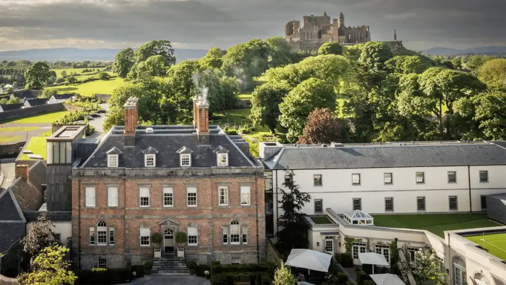 An elevated view of the historic red-brick Cashel Palace Hotel in Tipperary, featuring its 1730 Palladian architecture and the new Garden Wing, with the iconic Rock of Cashel situated on the hill in the background.