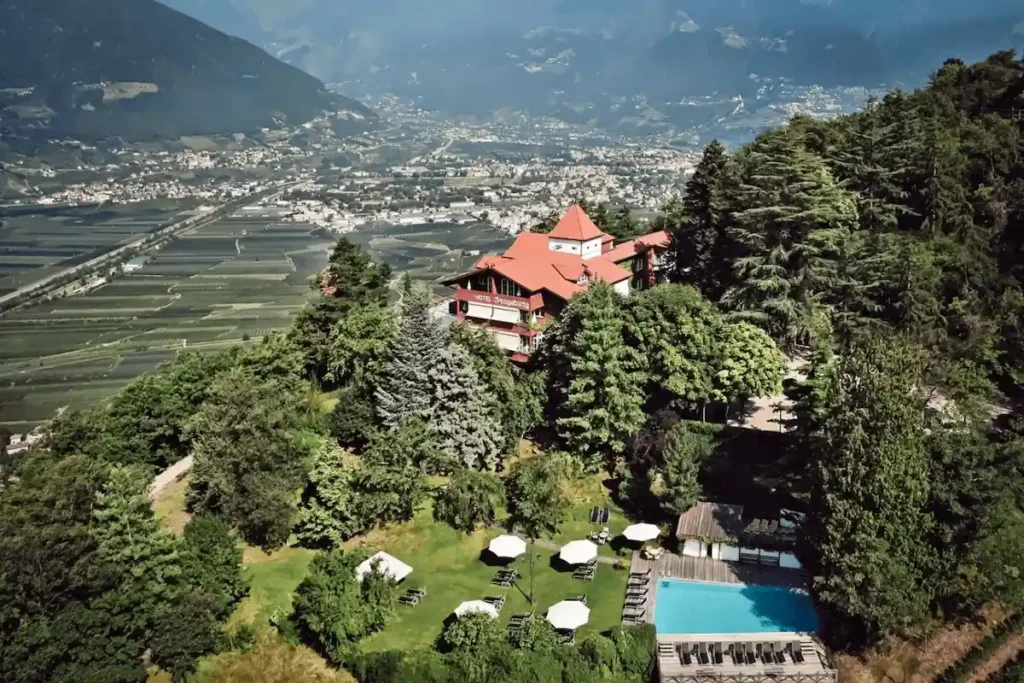 An elevated aerial view of the red-roofed Castel Fragsburg perched high on a wooded mountainside, featuring a sun-drenched pool deck and panoramic views over the terraced Adige Valley in South Tyrol.