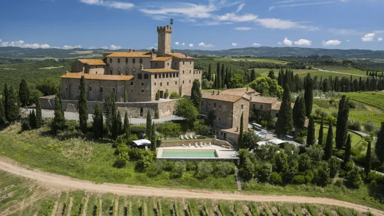 An elevated view of the medieval stone fortress of Castello Banfi il Borgo in Tuscany, featuring a prominent watchtower and terracotta roofs, surrounded by cypress trees and vast estate vineyards.