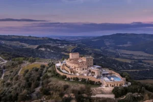 A wide twilight shot of the 11th-century Castello di Velona fortress on a hilltop in Val d'Orcia, featuring its restored stone watchtowers, expansive thermal pool decks, and panoramic views of the surrounding Tuscan landscape.
