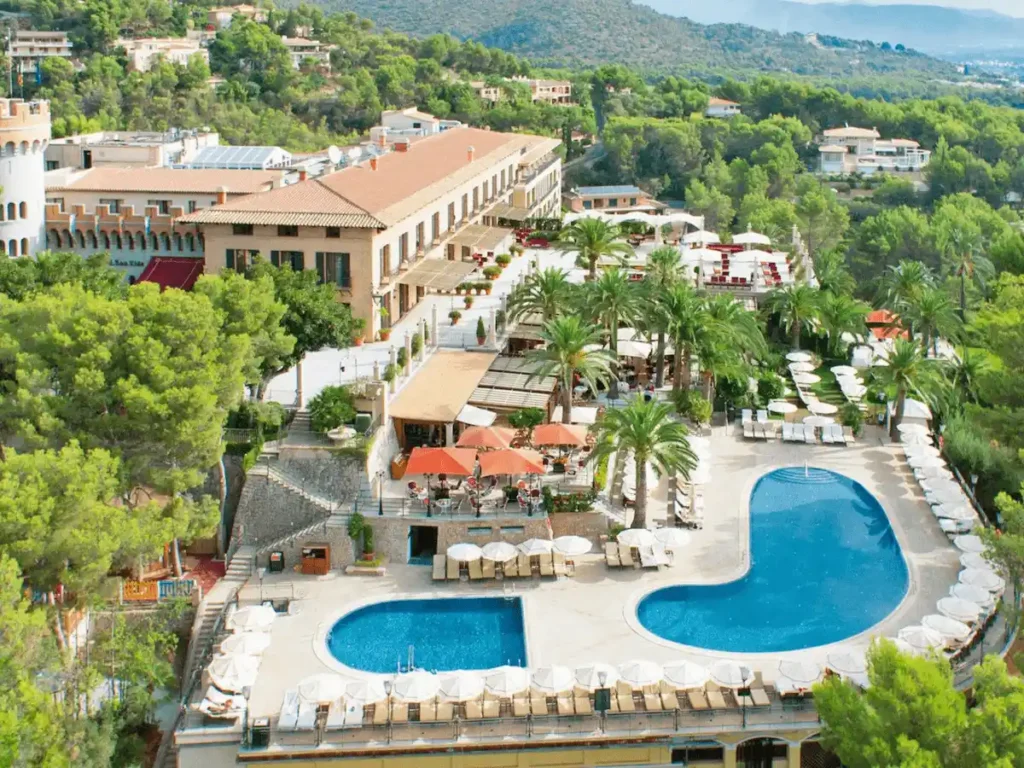 An elevated view of Castillo Hotel Son Vida in Mallorca, a 13th-century estate and former castle featuring traditional Mediterranean architecture, expansive sun terraces, and two turquoise swimming pools nestled within lush, pine-covered hills overlooking the Bay of Palma.