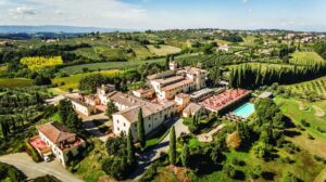 An elevated aerial view of the sprawling COMO Castello Del Nero estate in Tuscany, featuring terracotta-roofed villas, a large swimming pool, and manicured gardens surrounded by rolling vineyards and cypress trees.
