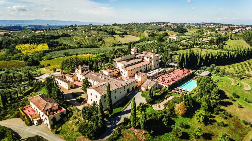An elevated aerial view of the sprawling COMO Castello Del Nero estate in Tuscany, featuring terracotta-roofed villas, a large swimming pool, and manicured gardens surrounded by rolling vineyards and cypress trees.