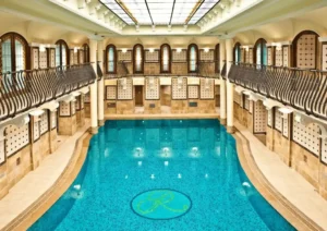 The historic Royal Spa at Corinthia Budapest featuring a grand 15-meter swimming pool surrounded by Art Deco stone pillars, intricate wall tiles, and an ornate stained-glass ceiling.