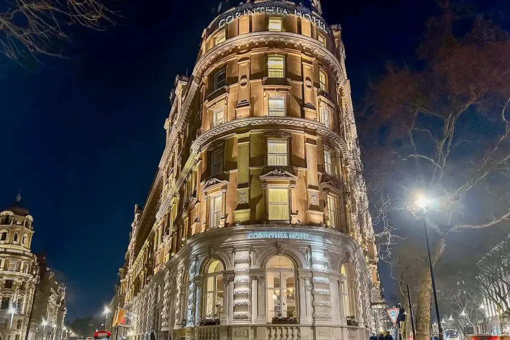 The illuminated Victorian facade of Corinthia London, a landmark 1885 building on Whitehall Place that formerly served as the Ministry of Defence, featuring an ornate curved corner, sand-colored stonework, and grand arched windows beneath a twilight sky.