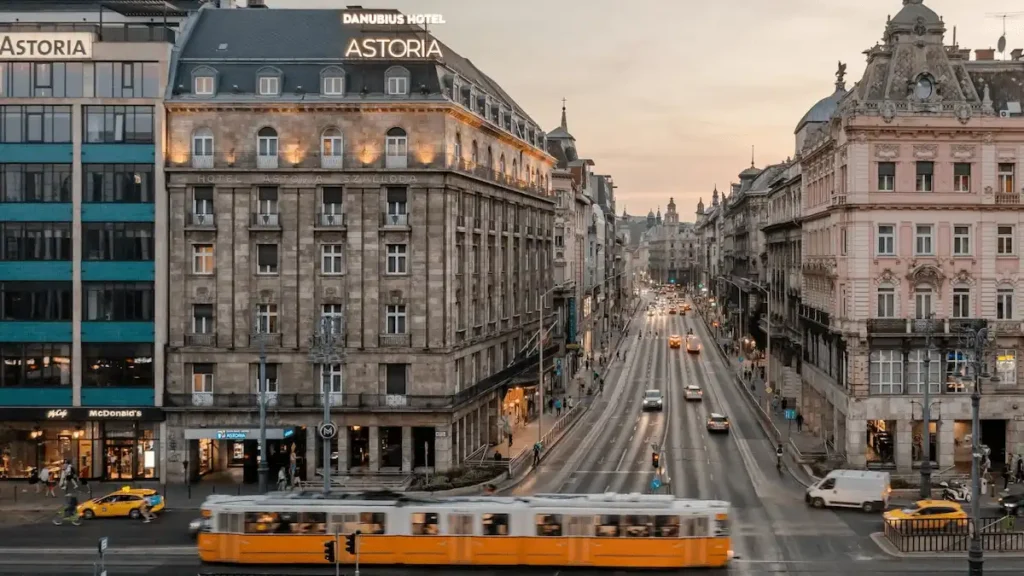 A high-angle dusk view of the historic stone facade of Danubius Hotel Astoria City Center, overlooking a busy Budapest street with a passing yellow tram and city lights.