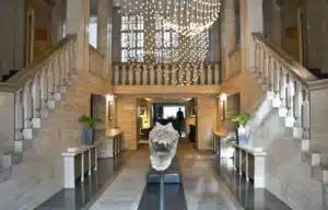 The grand 1930s travertine entrance hall of Das Stue Berlin, originally the Royal Danish Embassy, featuring dual sweeping staircases and a restored high-ceilinged interior designed by Johann Emil Schaudt.