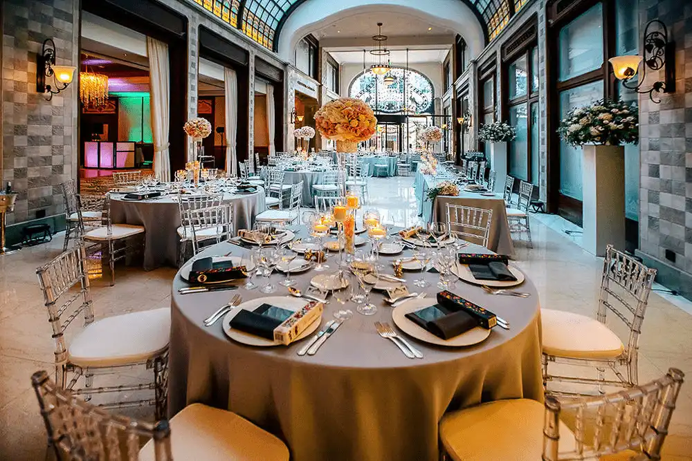 A formal banquet setup within the historic glass-roofed arcade of Four Seasons Gresham Palace Budapest, featuring mosaic tile floors and the iconic peacock ironwork gates in the distance.