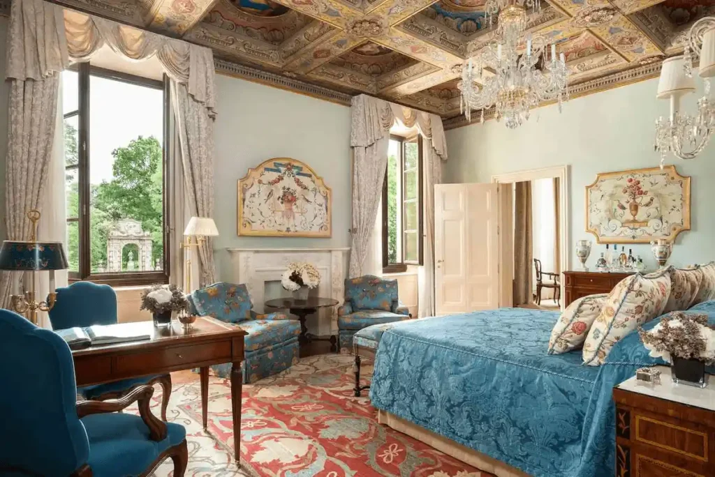 A lavish Noble Suite at Four Seasons Hotel Firenze, featuring an ornate coffered ceiling with gold detailing, historic frescoes, antique furniture, and large windows overlooking the Giardino della Gherardesca.