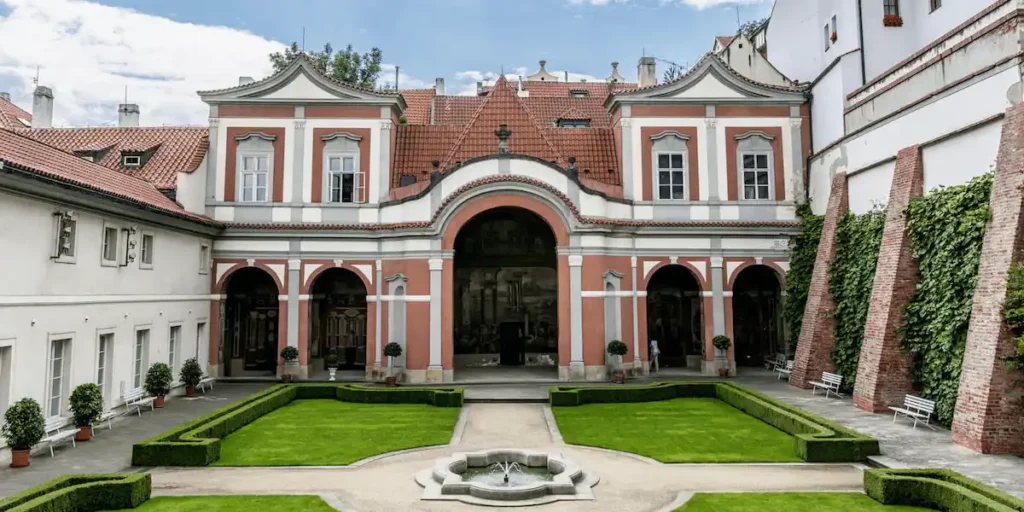 The ornate Renaissance courtyard of Golden Well Prague, featuring manicured boxwood hedges, a central stone fountain, and views of the historic red-tiled wings beneath Prague Castle.