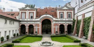 The ornate Renaissance courtyard of Golden Well Prague, featuring manicured boxwood hedges, a central stone fountain, and views of the historic red-tiled wings beneath Prague Castle.