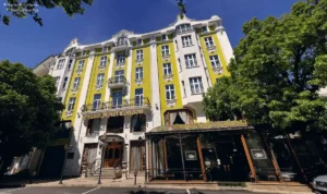 The vibrant yellow and white Art Nouveau facade of Grand Hotel London Varna, featuring ornate 1912 architectural carvings and the landmark street-level Grand Café terrace.