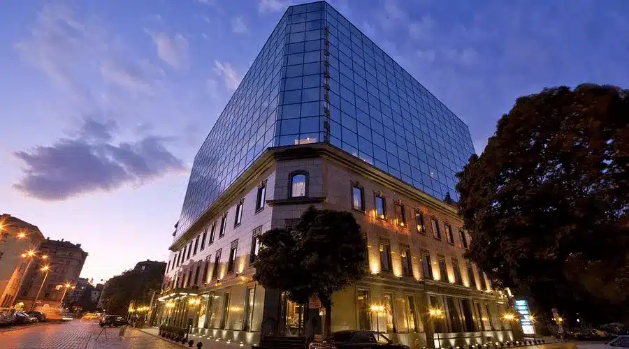 The contemporary stone and glass facade of Grand Hotel Sofia at twilight, overlooking the historic City Garden and the 19th-century architecture of the Sredets district.
