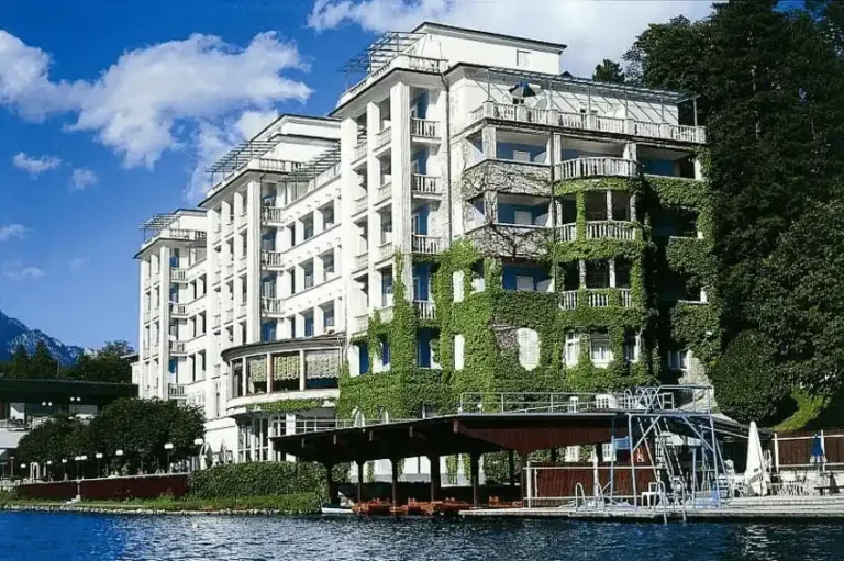 The white modernist facade of Grand Hotel Toplice covered in green ivy, situated directly on the shores of Lake Bled with a private wooden boathouse in the foreground.