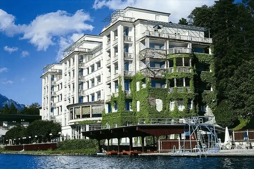The white modernist facade of Grand Hotel Toplice covered in green ivy, situated directly on the shores of Lake Bled with a private wooden boathouse in the foreground.
