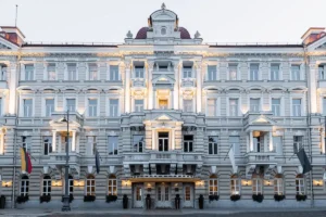 The symmetrical neo-classical facade of the Grand Hotel Vilnius at dusk, showcasing its grand white balconies, arched windows, and illuminated entrance facing Cathedral Square.