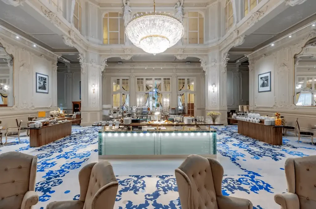 The magnificent Neo-Baroque Festive Hall of Grandhotel Pupp, featuring a massive crystal chandelier, ornate white stuccowork, and a gourmet breakfast buffet on a blue patterned carpet.