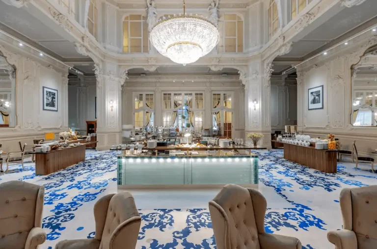 The magnificent Neo-Baroque Festive Hall of Grandhotel Pupp, featuring a massive crystal chandelier, ornate white stuccowork, and a gourmet breakfast buffet on a blue patterned carpet.