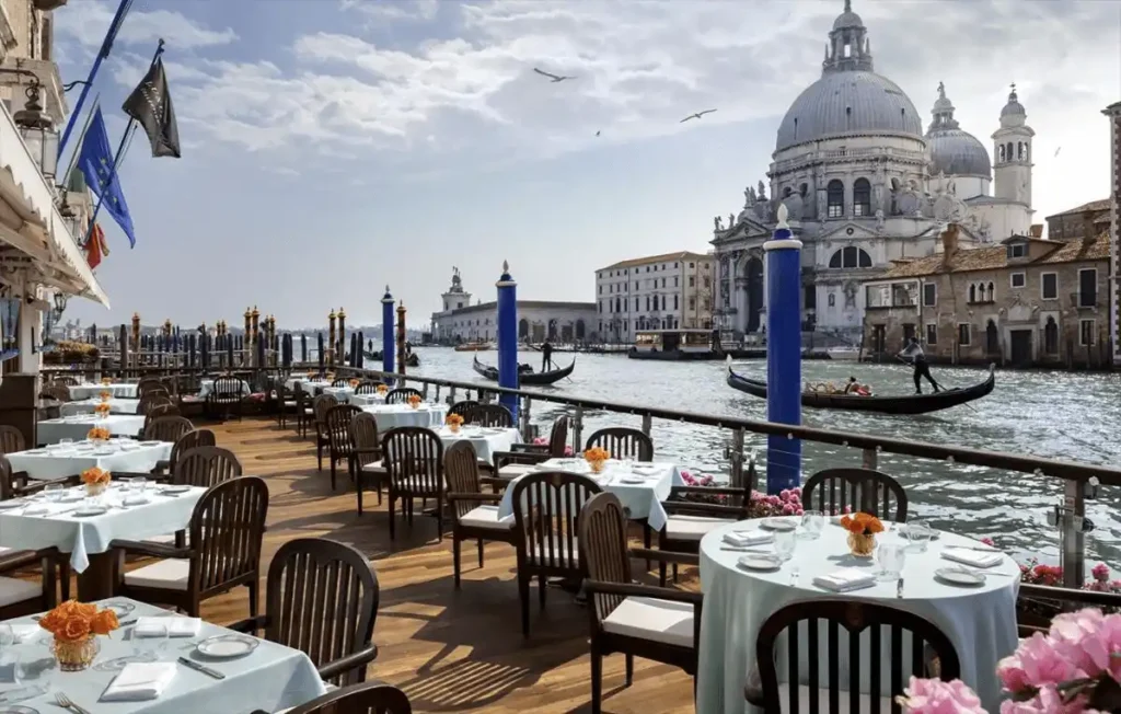 The famous Club del Doge terrace at the Gritti Palace Venice, a 15th-century noble residence, featuring outdoor dining tables and blue mooring poles overlooking the Grand Canal and the Santa Maria della Salute.