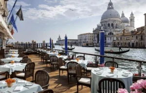 The famous Club del Doge terrace at the Gritti Palace Venice, a 15th-century noble residence, featuring outdoor dining tables and blue mooring poles overlooking the Grand Canal and the Santa Maria della Salute.