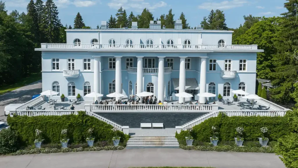 A symmetrical front view of the historic Hotel Haikko Manor & Spa in Porvoo, Finland, featuring its pale blue Neoclassical facade, grand white columns, and a wide sun terrace overlooking the coastal parkland.