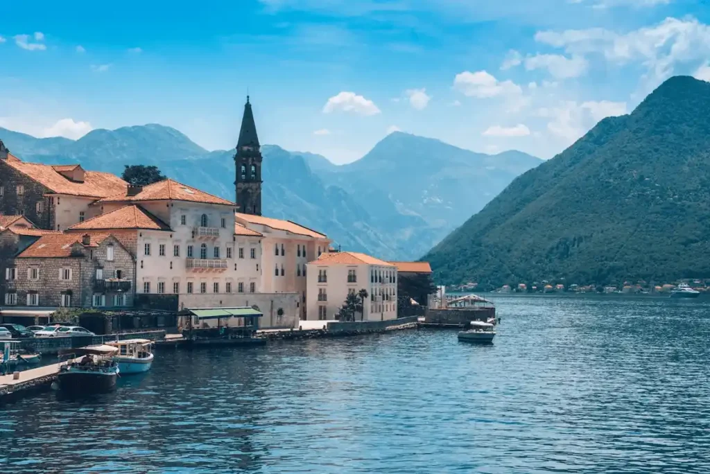 A stunning waterfront view of Heritage Grand Perast in Montenegro, featuring its historic stone palace architecture and bell tower against the backdrop of the Bay of Kotor mountains.