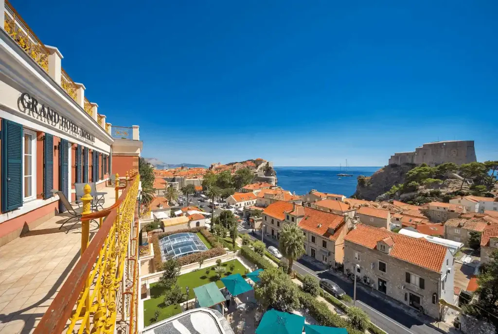 View from the yellow-railed balcony of Hilton Imperial Dubrovnik overlooking the terracotta roofs of the Old Town, the Adriatic Sea, and the historic Lovrijenac Fortress.