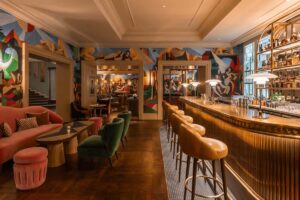 The interior of Bar Magritte at Hotel Amigo Brussels, featuring a long fluted brass bar, velvet cocktail seating in jewel tones, and expansive colorful wall murals inspired by the surrealist works of René Magritte.