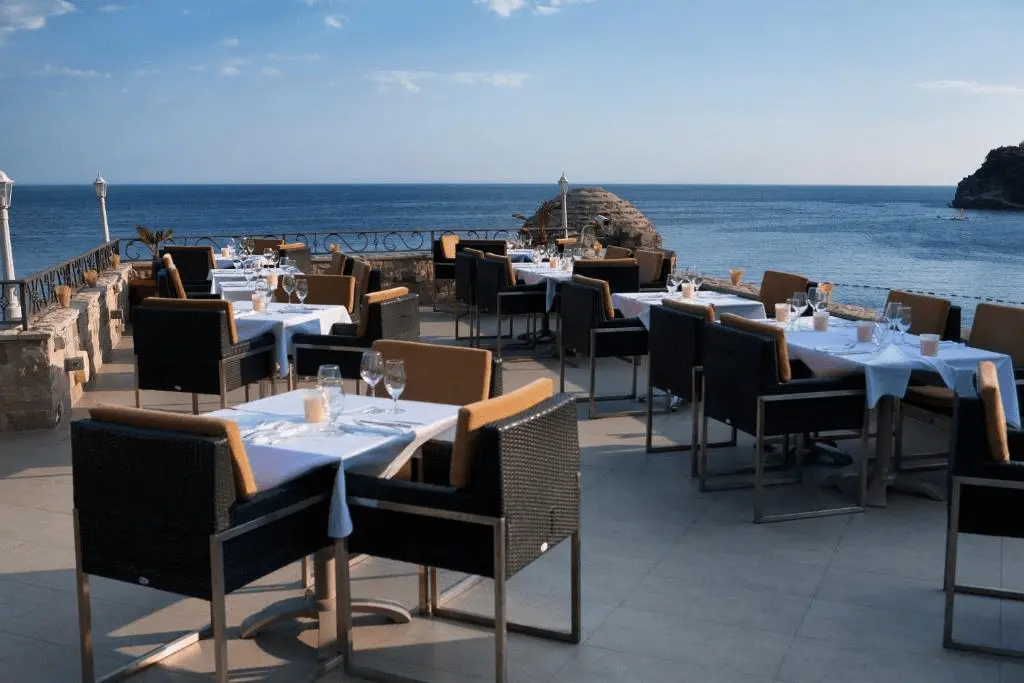 The rooftop restaurant terrace of Hotel Astoria Budva, featuring elegant table settings overlooking the medieval stone fortifications and the blue Adriatic Sea.