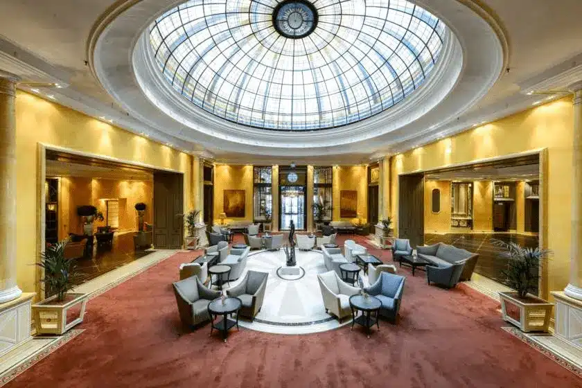 The expansive central Atrium lobby of Hotel Bayerischer Hof, featuring a magnificent stained-glass dome that dates back to the early 19th-century origins of the hotel, surrounded by upper-level galleries and luxury boutiques.