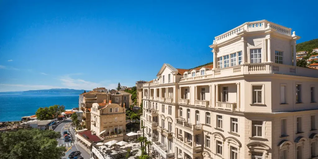 A high-angle view of the neo-baroque facade of Hotel Bellevue Opatija, situated along the coastal road with the blue Adriatic Sea and Kvarner Bay in the background.