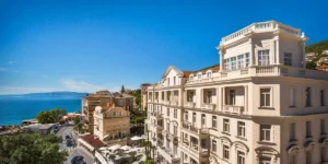 A high-angle view of the neo-baroque facade of Hotel Bellevue Opatija, situated along the coastal road with the blue Adriatic Sea and Kvarner Bay in the background.